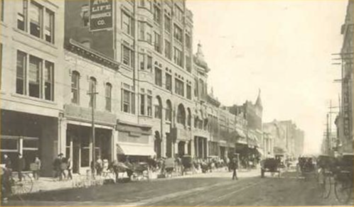 Historic street scene with horse-drawn carriages, buildings, and pedestrians in a bustling urban setting.
