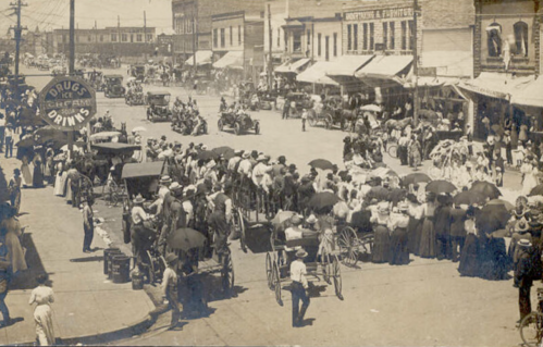 A historic street scene with crowds, vintage cars, and people in period clothing, shaded by umbrellas.