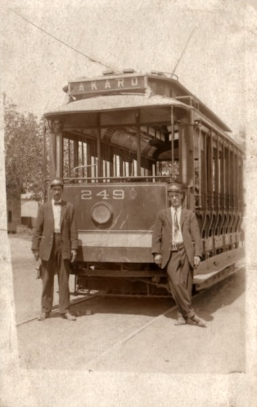 Two men in suits stand beside an old tram labeled "Akaroa," with a vintage sepia tone.