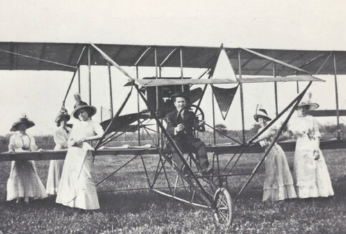A vintage photo of a man in an early airplane, surrounded by women in elegant dresses and large hats.