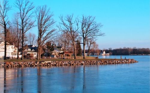 A serene lakeside view with bare trees, houses, and a calm blue sky reflecting on the water's surface.