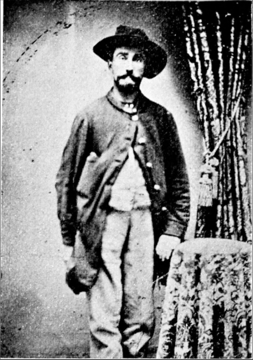 A historical black-and-white portrait of a man in a hat and coat, standing beside a table with a patterned cloth.