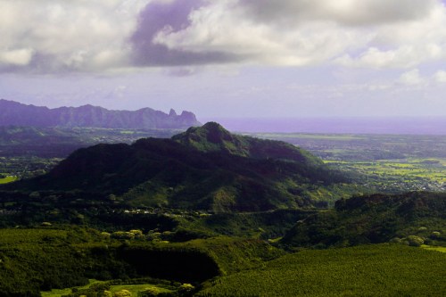 A panoramic view of lush green mountains and valleys under a cloudy sky, with distant peaks visible on the horizon.