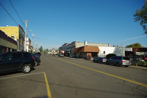 A quiet street scene with parked cars and small businesses under a clear blue sky.