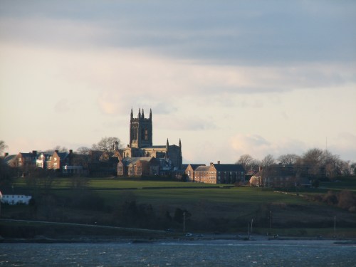 A scenic view of a town with a prominent church tower, surrounded by green fields and a cloudy sky.