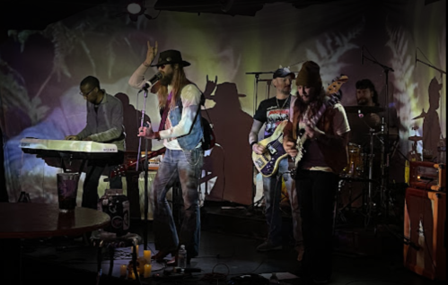 A band performs on stage with colorful projections in the background, featuring a keyboardist, guitarist, and drummer.