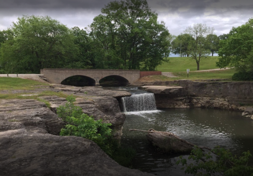 A serene landscape featuring a small waterfall flowing into a calm river, with a stone bridge and lush greenery in the background.