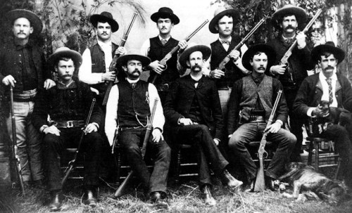 A historic black-and-white photo of ten men in Western attire, armed with rifles, posing together outdoors.