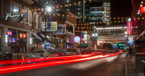 A vibrant city street at night, lined with illuminated shops and restaurants, with light trails from passing cars.