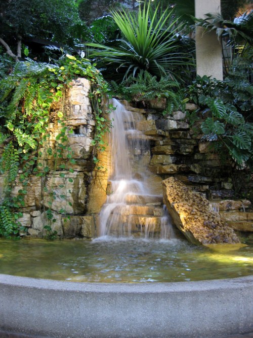 A serene waterfall cascades over rocks into a tranquil pool, surrounded by lush greenery and tropical plants.