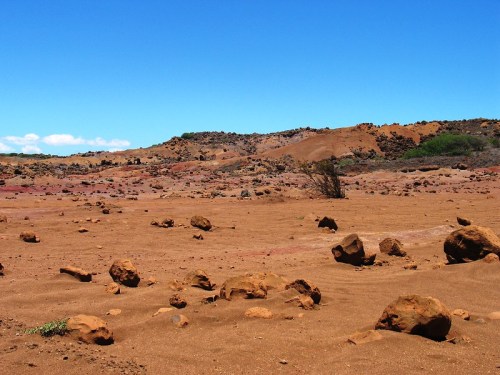 A barren landscape with reddish-brown soil, scattered rocks, and sparse vegetation under a clear blue sky.