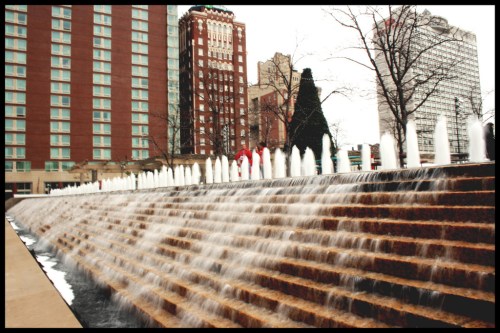 A cascading fountain flows down steps, with buildings and a tree in the background on a cloudy day.
