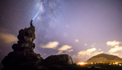 A person stands on a rock, shining a flashlight at the starry sky, with mountains and lights in the background.