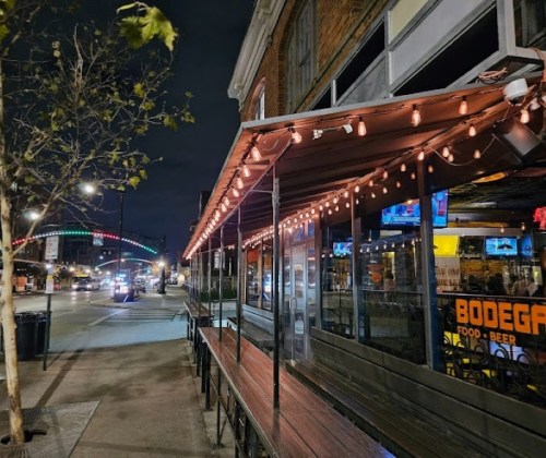 A dimly lit outdoor seating area of a bar, adorned with string lights, along a city street at night.