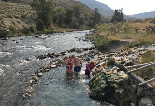 A group of people wade in a river surrounded by rocky banks and greenery, with mountains in the background.