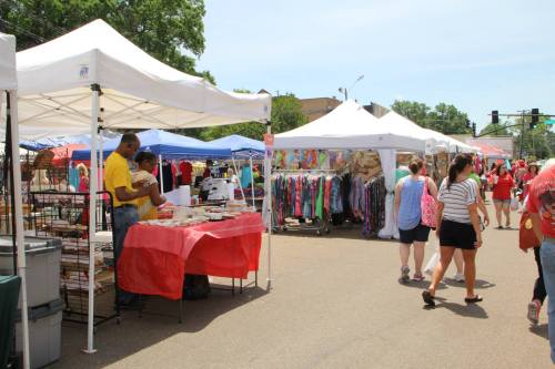 A vibrant outdoor market with colorful tents, vendors, and shoppers browsing various goods on a sunny day.