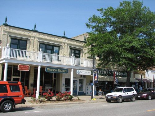 A street view of a charming town with shops, including Watson Realty and a hardware store, under a clear blue sky.