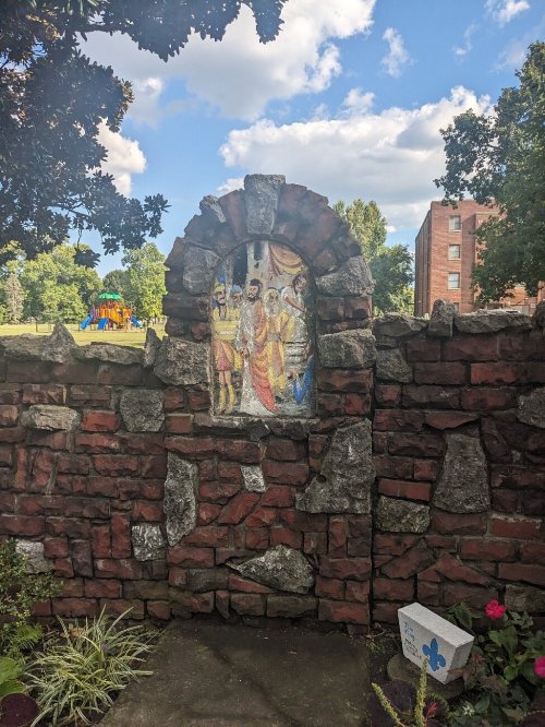 A stone wall with a colorful mural of figures, set against a blue sky and green trees in a park-like area.
