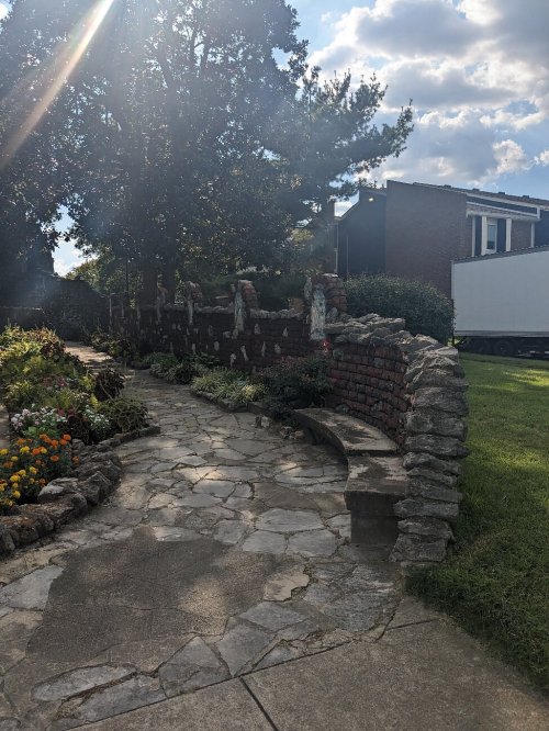 A stone pathway lined with flowers leads to a brick wall and a large tree under a sunny sky.