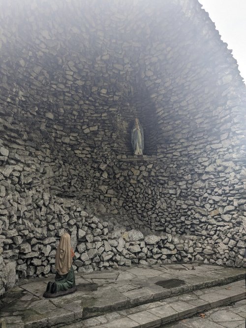 A person kneels in prayer before a stone altar featuring a statue of a figure, surrounded by a rocky archway.