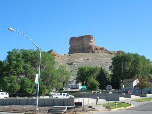 A rocky butte rises above a small town, surrounded by trees and buildings under a clear blue sky.