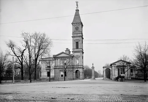 Historic black-and-white photo of a clock tower and buildings on a street, with bare trees and an early automobile.