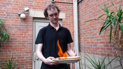 A person stands outside, holding a plate with flames rising from it, surrounded by brick walls and plants.