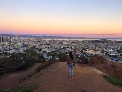 A person stands on a hilltop overlooking a city at sunset, with vibrant colors in the sky and a view of the coastline.