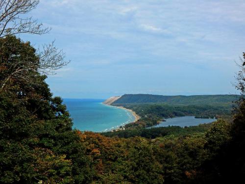 A scenic view of a coastline with sandy cliffs, lush greenery, and a calm blue lake under a partly cloudy sky.