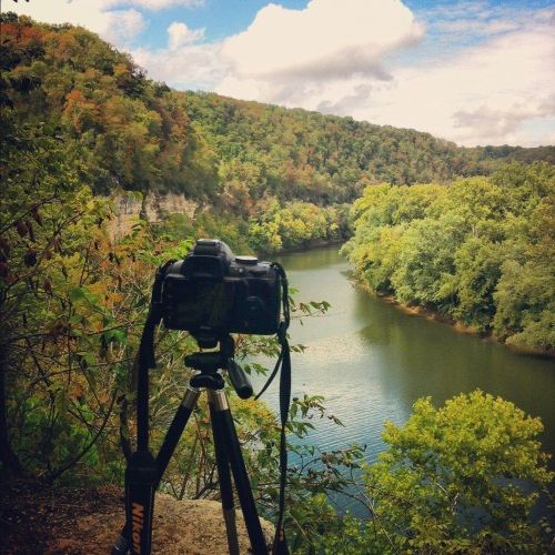 A camera on a tripod overlooking a serene river surrounded by lush green trees and rocky cliffs under a cloudy sky.