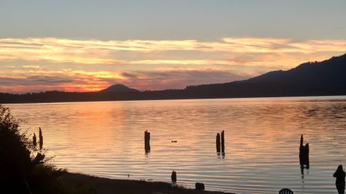 Sunset over a calm lake, with silhouettes of trees and wooden posts reflected in the water, and mountains in the background.