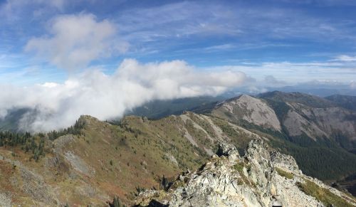 A panoramic view of rugged mountains under a blue sky with scattered clouds and lush green valleys below.