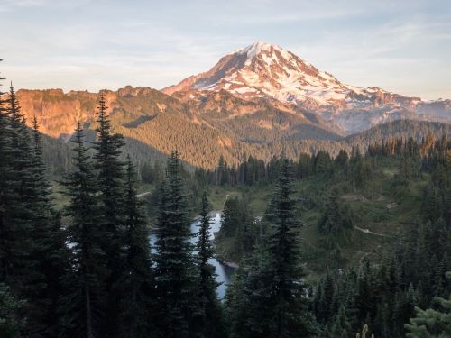 Majestic mountain landscape with snow-capped peak, lush green forests, and a serene river in the foreground.