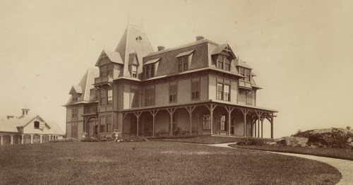 A historic large Victorian-style house with multiple gables, set on a grassy hill with a winding path.