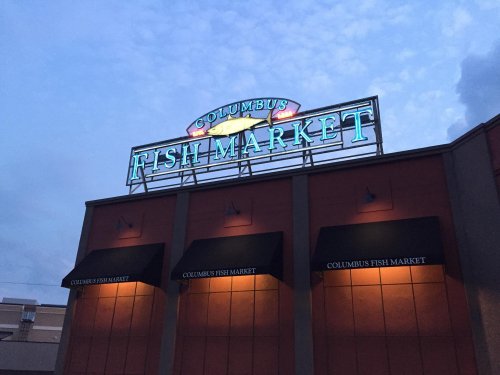 Sign for Columbus Fish Market illuminated at dusk, featuring a colorful logo and awnings.