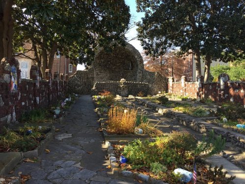 A peaceful cemetery path lined with stone walls and overgrown gardens, surrounded by trees and sunlight.