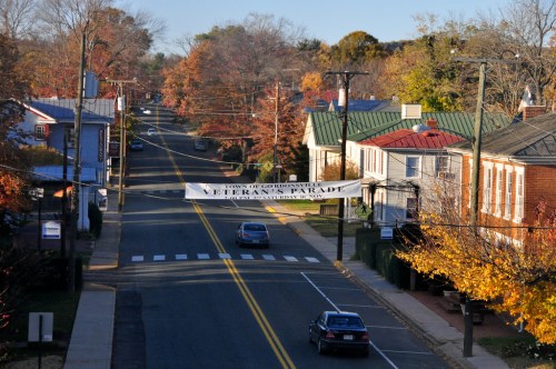 A street lined with trees in autumn colors, featuring a banner for a Veterans Parade across the road.