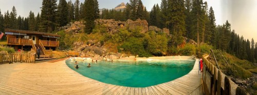 A scenic outdoor pool surrounded by trees and rocks, with people swimming and a lodge in the background.