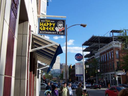 Sign for "The Happy Greek Restaurant & Pub" on a city street with pedestrians and buildings in the background.