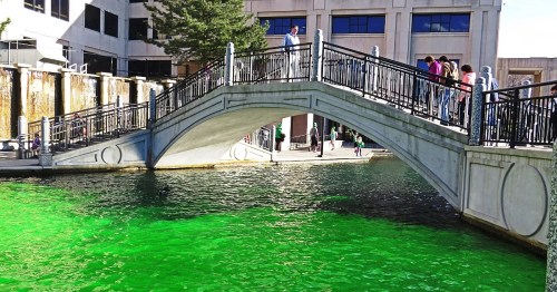 A bridge over vibrant green water, with people walking along the path and buildings in the background.