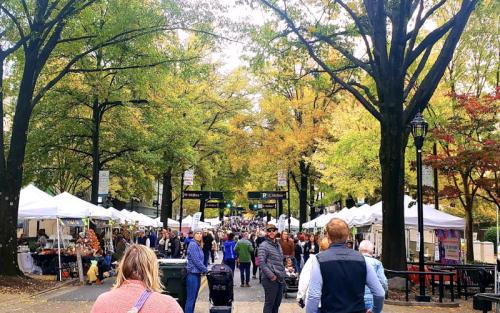 A bustling outdoor market lined with white tents, surrounded by colorful autumn trees and a crowd of people.