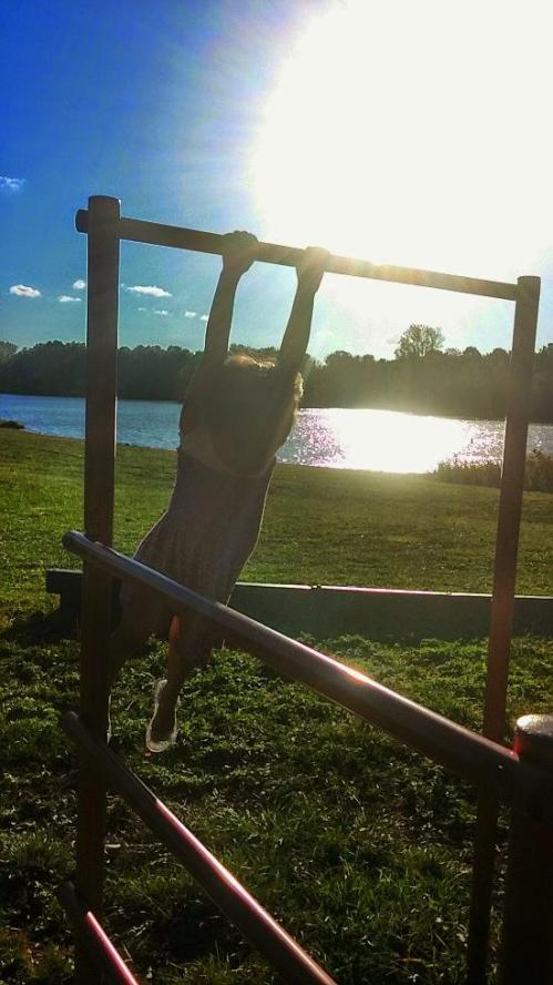 A child hangs from a playground bar with a lake and sunlit sky in the background.