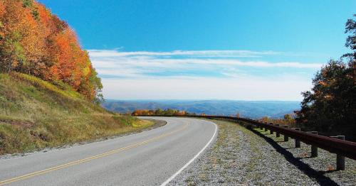 A winding road through vibrant autumn foliage under a clear blue sky, with rolling hills in the background.