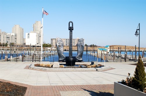 A large anchor sculpture in a waterfront park, with a marina and buildings in the background under a clear blue sky.