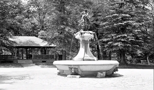 A vintage black-and-white photo of a fountain surrounded by trees, with a gazebo in the background.