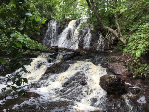 A serene waterfall cascades over rocks, surrounded by lush green trees and foliage in a tranquil forest setting.