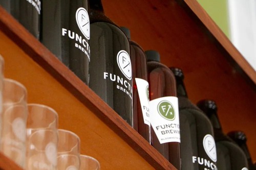 Bottles of Function Brewing displayed on a wooden shelf, with clear glasses in the foreground.