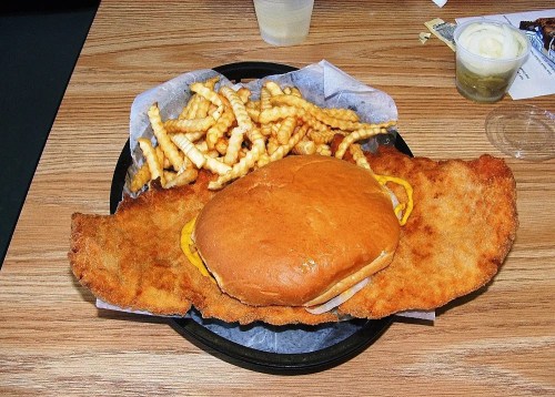 A large breaded fried cutlet with a cheeseburger on top, served with crinkle-cut fries on a black tray.