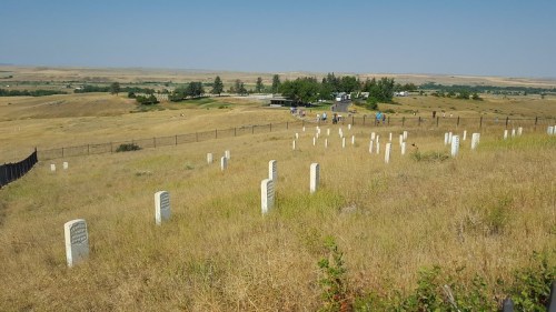 A wide view of a grassy cemetery with white headstones, set against a backdrop of rolling hills and a distant gathering.