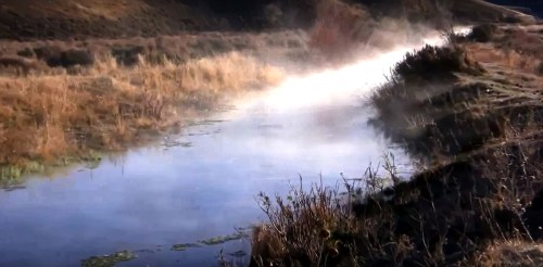 A misty river flows through a grassy landscape, with steam rising in the cool morning air.